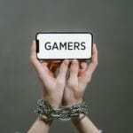Dłonie skutego gracza trzymającego smartfon z napisem GAMERS